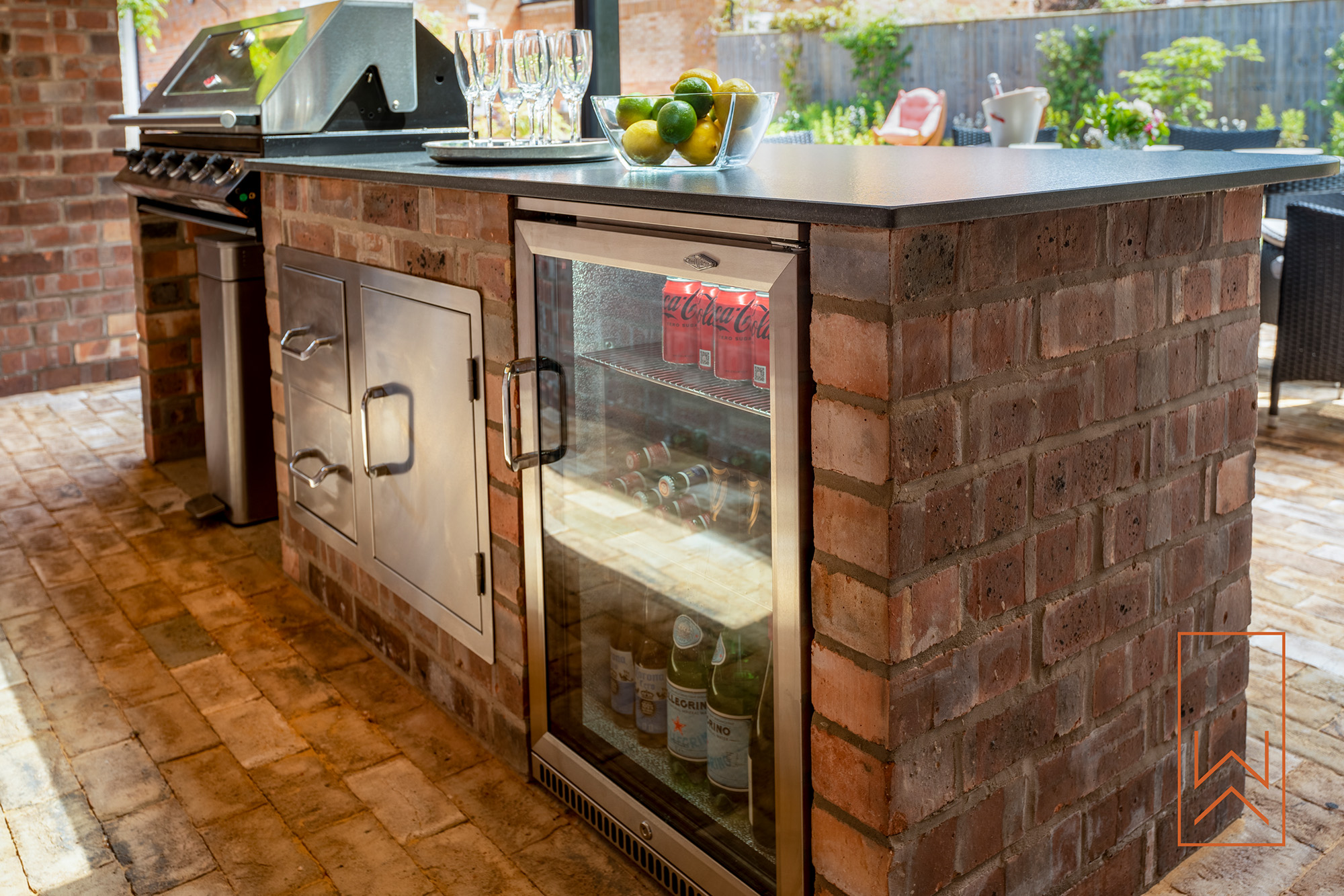 Brick outdoor kitchen with integrated fridge and barbecue in landscaped garden.
